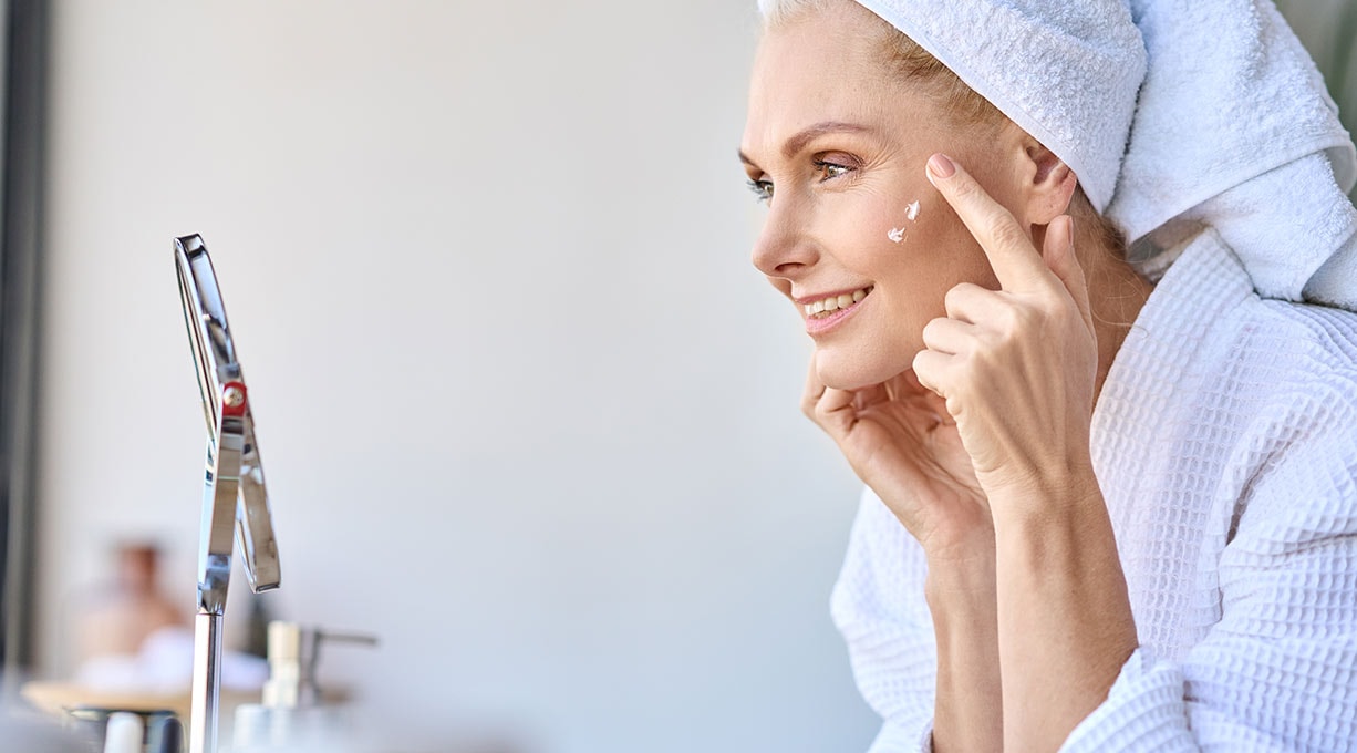 Eine Frau mittleren Alters mit Handtuchturban auf dem Kopf tupft sich mit dem Zeigefinger Gesichtspflege mit Kollagen-Peptiden auf die Haut