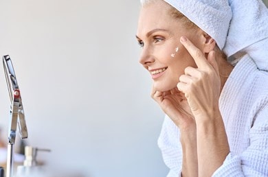 Eine Frau mittleren Alters mit Handtuchturban auf dem Kopf tupft sich mit dem Zeigefinger Gesichtspflege mit Kollagen-Peptiden auf die Haut