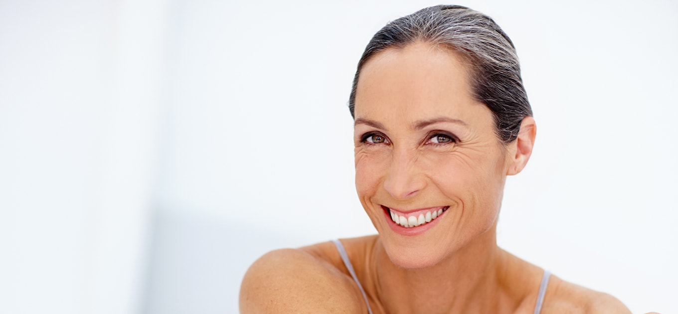 Lachende Frau mittleren Alters mit Mimikfalten und zurückgebundenen Haaren vor hellem Hintergrund