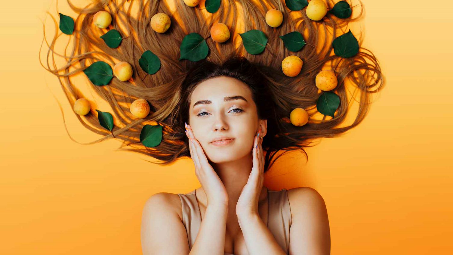 Frau liegt auf orangenem Untergrund, ihre langen Haare sind von Aprikosen bedeckt