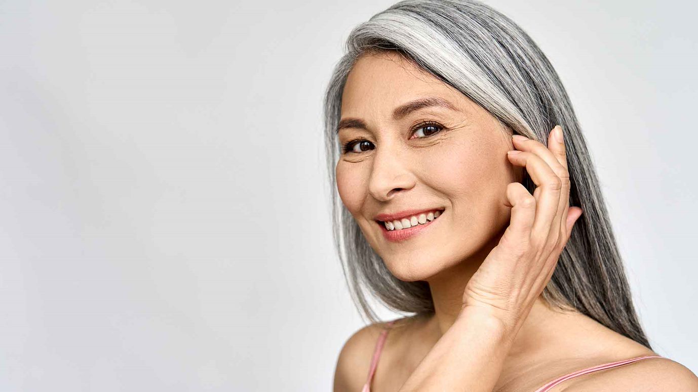 Ältere Frau mit graunen langen Haaren lächelt während sie ihr Gesicht sanft berührt