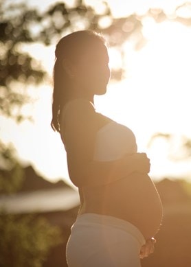 Eine schwangere Frau steht draussen während sie von der Sonne im Hintergrund angestrahlt wird