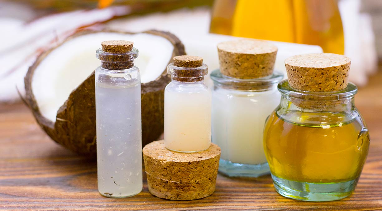 Mehrere kleine Glasflaschen mit Korkverschluss, gefüllt mit verschiedenen Ölen und Flüssigkeiten, neben einer aufgeschnittenen Kokosnuss.