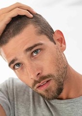Ein Mann betrachtet sein Haar nachdenklich im Spiegel und hebt dabei eine Hand an den Kopf.