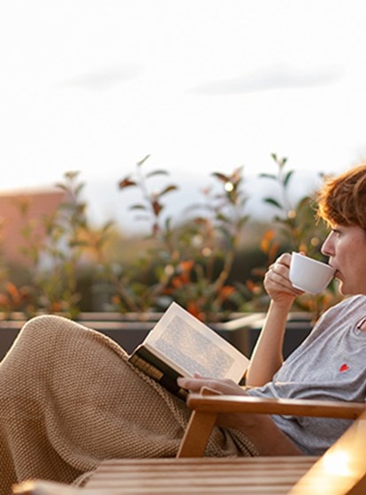 Frau entspannt auf der Terrasse, liest ein Buch und trinkt Kaffee bei Sonnenuntergang