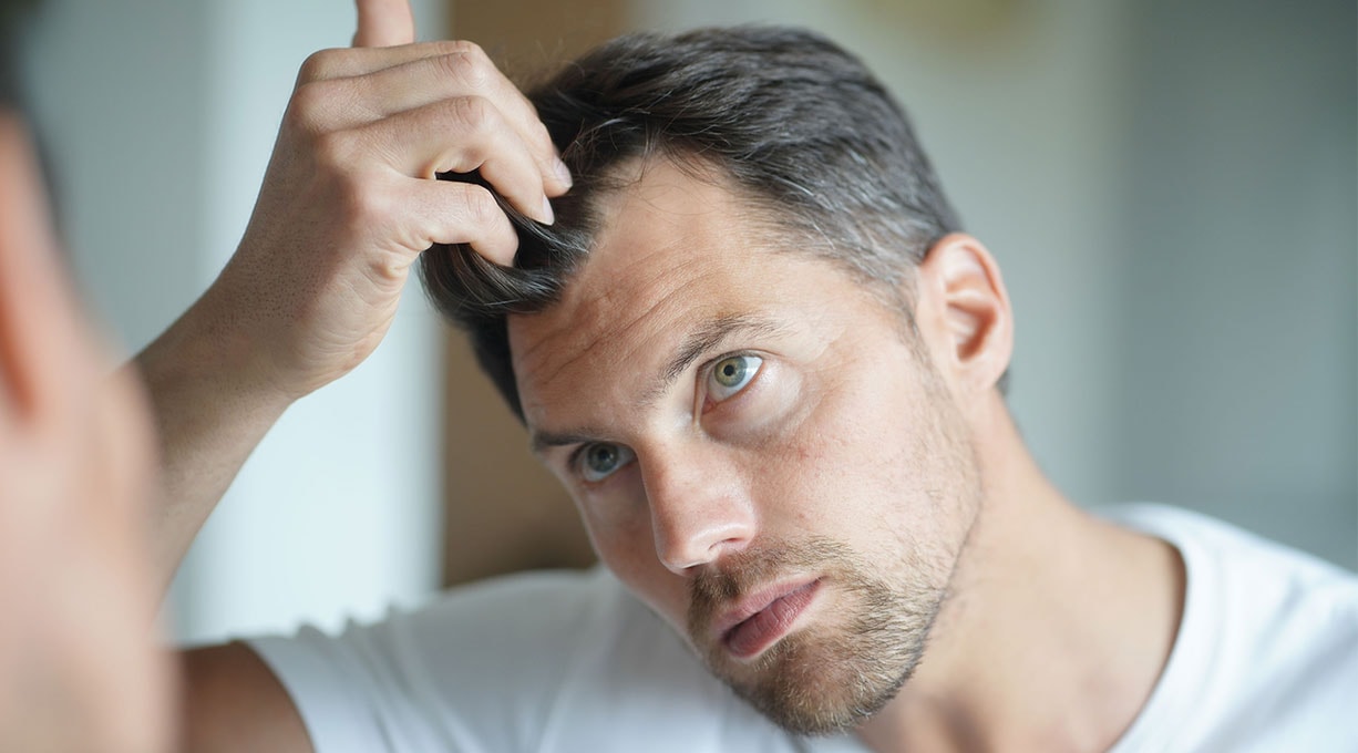 Eine Person in weißem Shirt untersucht aufmerksam ihr Haar, indem sie es mit der Hand anhebt. Die Szene spielt in einem Innenraum.