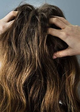 Eine Person greift sich mit beiden Händen in ihr langes, braunes, leicht zerzaustes Haar, Hintergrund ist unscharf und grau-blau.