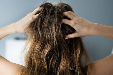 Eine Person greift sich mit beiden Händen in ihr langes, braunes, leicht zerzaustes Haar, Hintergrund ist unscharf und grau-blau.