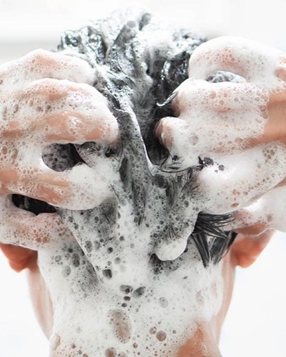 Person wäscht Haare unter der Dusche, die Hände massieren Schaum ins nasse Haar, Nahaufnahme des Kopfs von hinten.