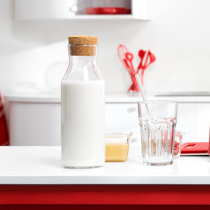 Glasflasche mit Milch neben Trinkglas und kleinem Behälter auf einer weißen Küchenarbeitsplatte
