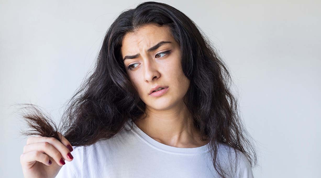 Person mit langen, dunklen, welligen Haaren, die mit der Hand eine Haarsträhne hält. Sie trägt ein weißes Oberteil vor neutralem Hintergrund.