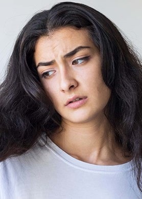 Person mit langen, dunklen, welligen Haaren, die mit der Hand eine Haarsträhne hält. Sie trägt ein weißes Oberteil vor neutralem Hintergrund.