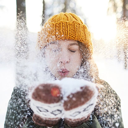 Person mit gelber Mütze pustet Schnee aus den Handschuhen im winterlichen Wald