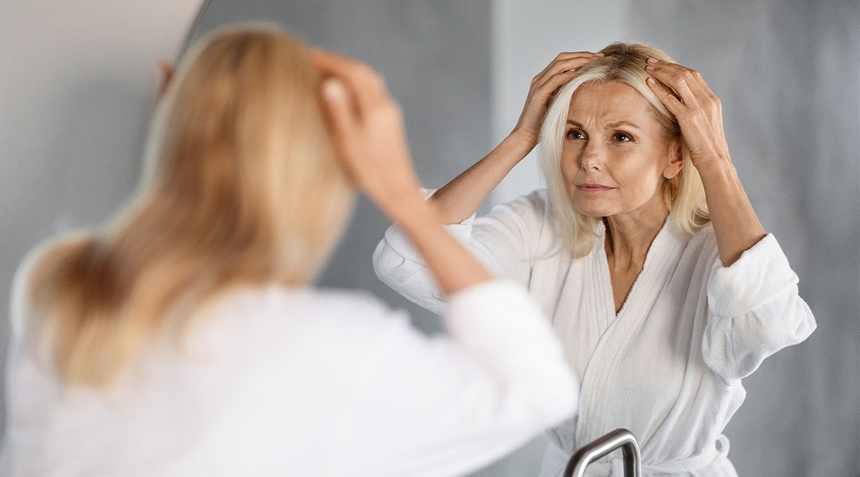 Frau mittleren Alters vor dem Spiegel begutachtet ihren Scheitel