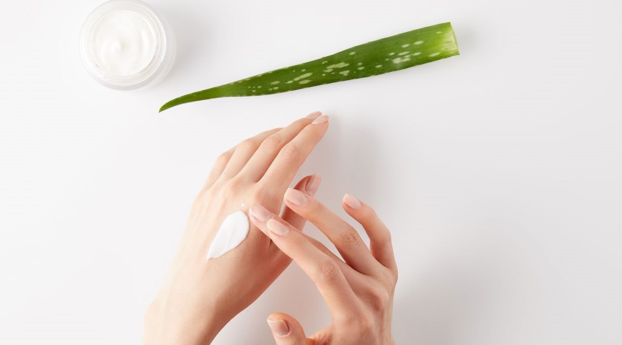 Ein Aloe vera Blatt, ein Tiegel Creme und zwei Hände, von denen eine Creme aufträgt