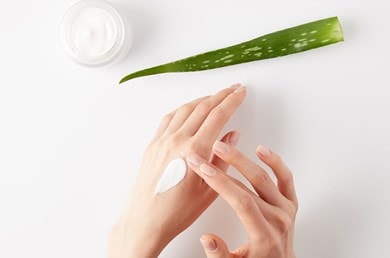 Ein Aloe vera Blatt, ein Tiegel Creme und zwei Hände, von denen eine Creme aufträgt