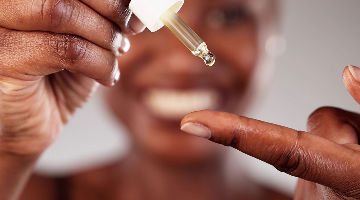 Eine Frau gibt sich mit einer Pipette Retinol-Serum auf dem Finger; im Hintergrund ist ihr unscharfes Lächeln erkennbar