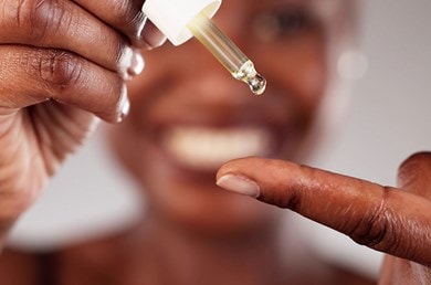 Eine Frau gibt sich mit einer Pipette Retinol-Serum auf dem Finger; im Hintergrund ist ihr unscharfes Lächeln erkennbar