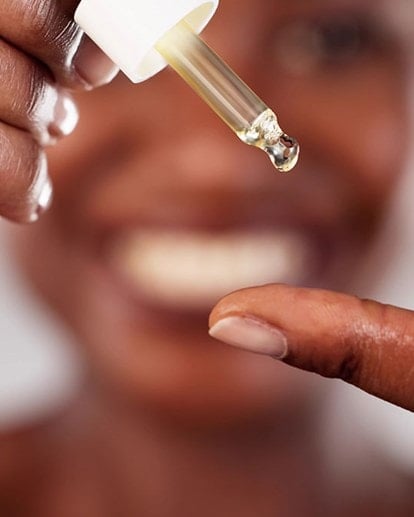 Eine Frau gibt sich mit einer Pipette Retinol-Serum auf dem Finger; im Hintergrund ist ihr unscharfes Lächeln erkennbar