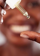 Eine Frau gibt sich mit einer Pipette Retinol-Serum auf dem Finger; im Hintergrund ist ihr unscharfes Lächeln erkennbar