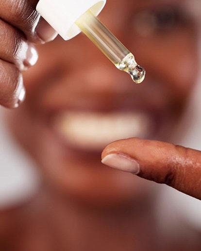 Eine Frau gibt sich mit einer Pipette Retinol-Serum auf dem Finger; im Hintergrund ist ihr unscharfes Lächeln erkennbar