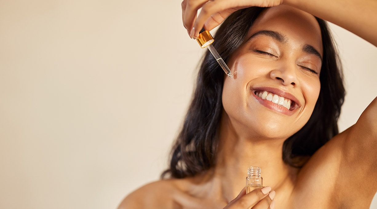 Eine lächelnde Frau trägt sich Serum mit einer Pipette auf die Wange auf