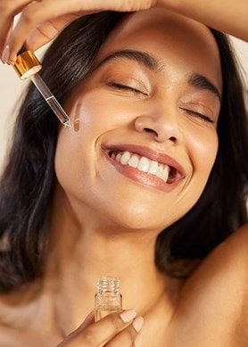 Eine lächelnde Frau trägt sich Serum mit einer Pipette auf die Wange auf