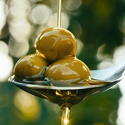 Mehrere grüne Oliven auf einem Löffel, über die Olivenöl läuft