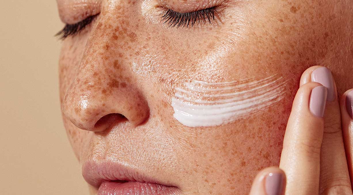 Frau mit geschlossenen Augen und Sommersprossen gibt Vitamin-C-Creme auf die Wange