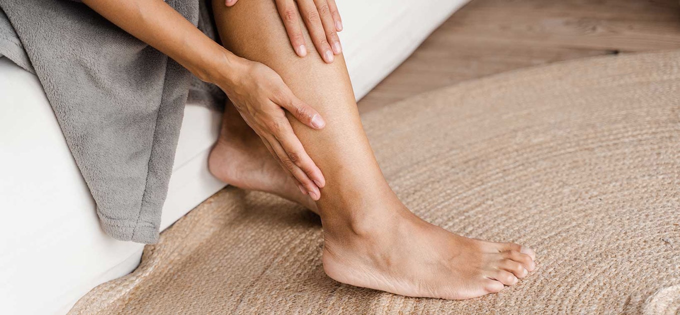 Frau sitzt auf einer Couch, trägt einen grauen Bademantel und hält ihre Hände auf ihrem Unterschenkel, Fokus auf die Haut der Beine in Bezug auf das Thema Hyperpigmentierung.