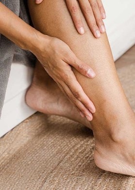 Frau sitzt auf einer Couch, trägt einen grauen Bademantel und hält ihre Hände auf ihrem Unterschenkel, Fokus auf die Haut der Beine in Bezug auf das Thema Hyperpigmentierung.