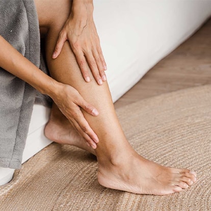 Frau sitzt auf einer Couch, trägt einen grauen Bademantel und hält ihre Hände auf ihrem Unterschenkel, Fokus auf die Haut der Beine in Bezug auf das Thema Hyperpigmentierung.