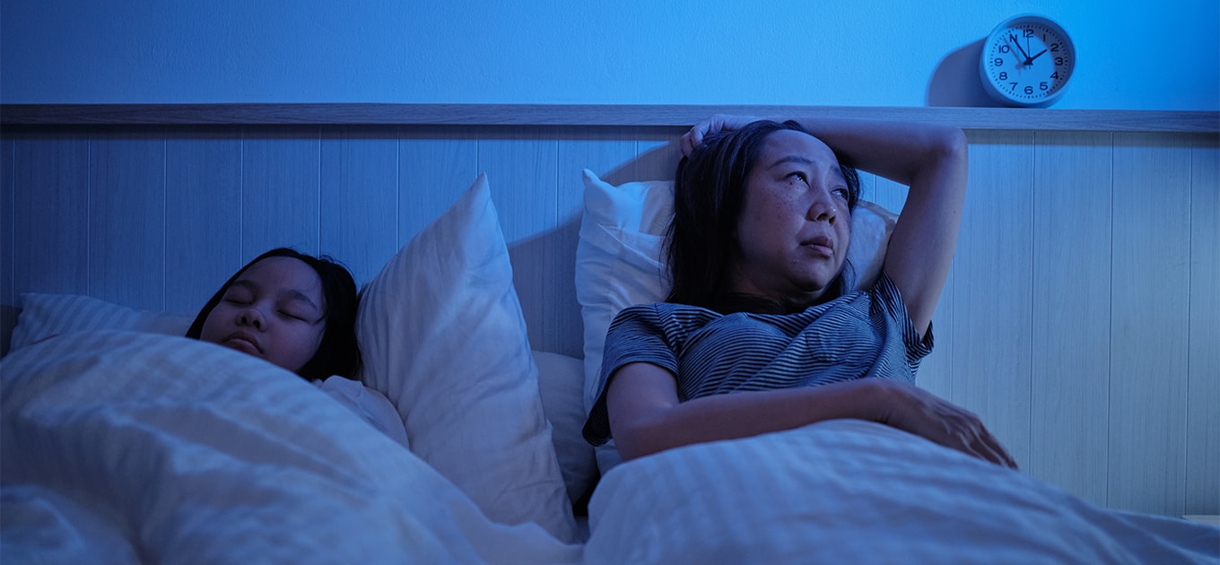 Frau liegt nachts wach im Bett wegen Nachtschweiß neben einem schlafenden Kind