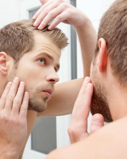 Junger Mann betrachtet im Spiegel kritisch seine Haare und Kopfhaut auf Anzeichen von Haarausfall.