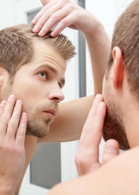 Junger Mann betrachtet im Spiegel kritisch seine Haare und Kopfhaut auf Anzeichen von Haarausfall.
