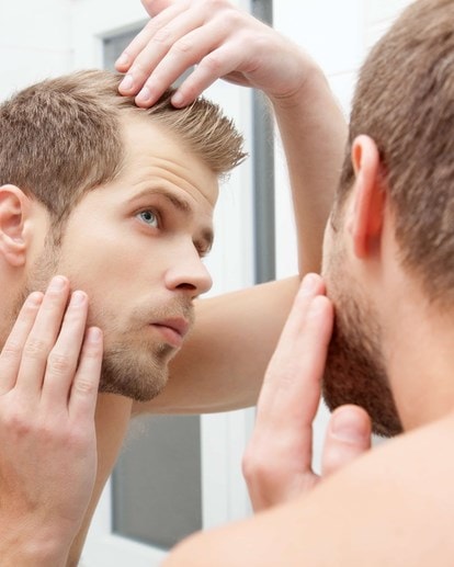 Junger Mann betrachtet im Spiegel kritisch seine Haare und Kopfhaut auf Anzeichen von Haarausfall.