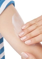 Eine Hand trägt weiße Creme auf den Arm einer Person mit blau-weiß gestreiftem Shirt auf, um die Haut zu pflegen und Feuchtigkeit zu spenden.