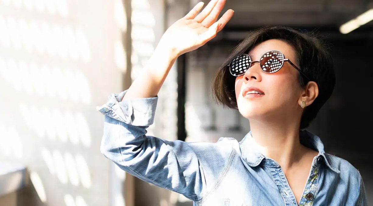 Person in Jeanshemd hebt eine Hand in hellem, sonnenbeschienenem Raum. Licht- und Schattenmuster fallen an der Wand entlang.