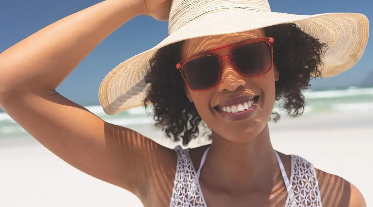 Person mit lockigem Haar trägt einen Strohhut und steht am sonnigen Strand, im Hintergrund das Meer und blauer Himmel.