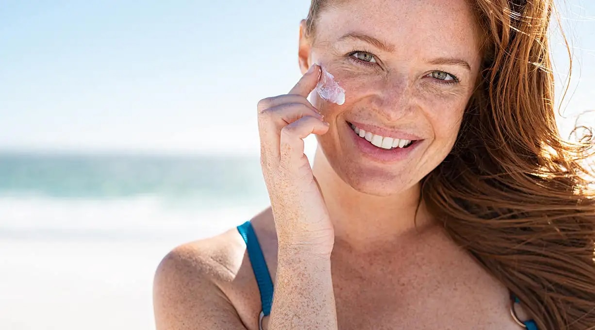 Person in blauem Oberteil am Strand mit Sonnenschein, braunen Haaren und Sommersprossen. Im Hintergrund Meer und blauer Himmel.