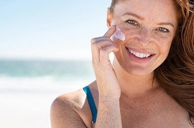 Person in blauem Oberteil am Strand mit Sonnenschein, braunen Haaren und Sommersprossen. Im Hintergrund Meer und blauer Himmel.