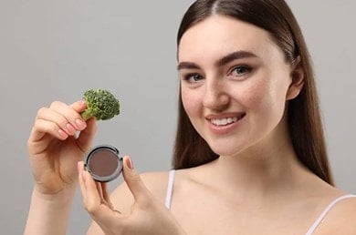 Eine Person hält ein Stück Brokkoli in einer Hand und ein rundes Make-up-Produkt in der anderen vor einem grauen Hintergrund.