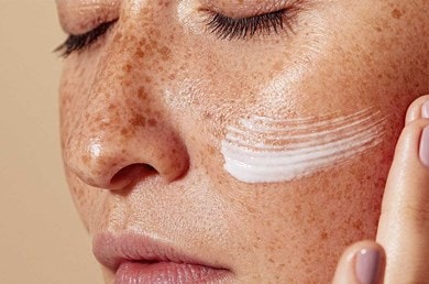 Nahaufnahme einer Frau mit Sommersprossen vor beigefarbenen Hintergrund. Auf der rechten Wange sieht man eine Creme die gerade aufgetragen wurde.
