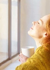 Eine Frau sitzt mit geschlossenen Augen und einer Tasse vor einem geschlossenen Fenster und lässt die Sonne in ihr Gesicht scheinen