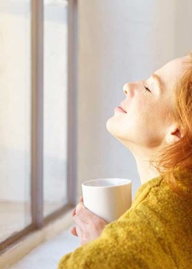 Eine Frau sitzt mit geschlossenen Augen und einer Tasse vor einem geschlossenen Fenster und lässt die Sonne in ihr Gesicht scheinen