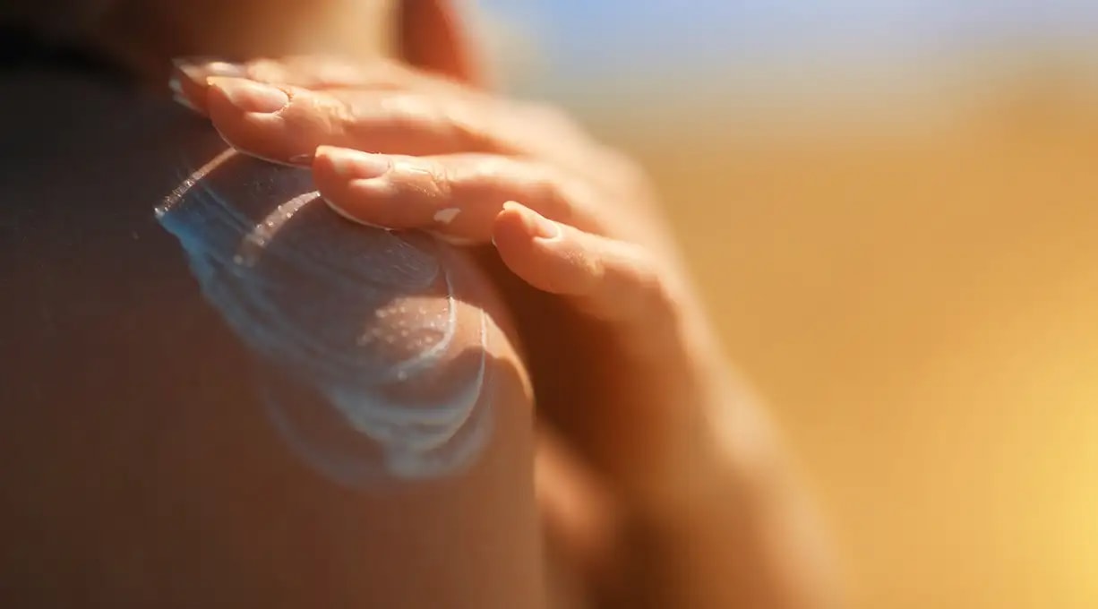 Hand trägt Sonnencreme auf nackter Haut auf, im Hintergrund unscharfer, sonniger Strand. Schutz vor Sonne und Pflege der Haut.