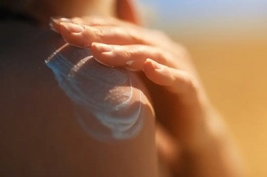 Hand trägt Sonnencreme auf nackter Haut auf, im Hintergrund unscharfer, sonniger Strand. Schutz vor Sonne und Pflege der Haut.