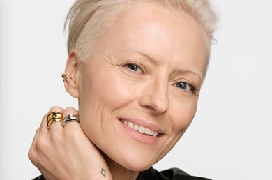 Person mit kurzem, blondem Haar, schwarzer Lederjacke und mehreren goldenen und silbernen Ringen an der Hand vor neutralem Hintergrund.
