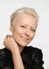 Person mit kurzem, blondem Haar, schwarzer Lederjacke und mehreren goldenen und silbernen Ringen an der Hand vor neutralem Hintergrund.