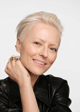 Person mit kurzem, blondem Haar, schwarzer Lederjacke und mehreren goldenen und silbernen Ringen an der Hand vor neutralem Hintergrund.